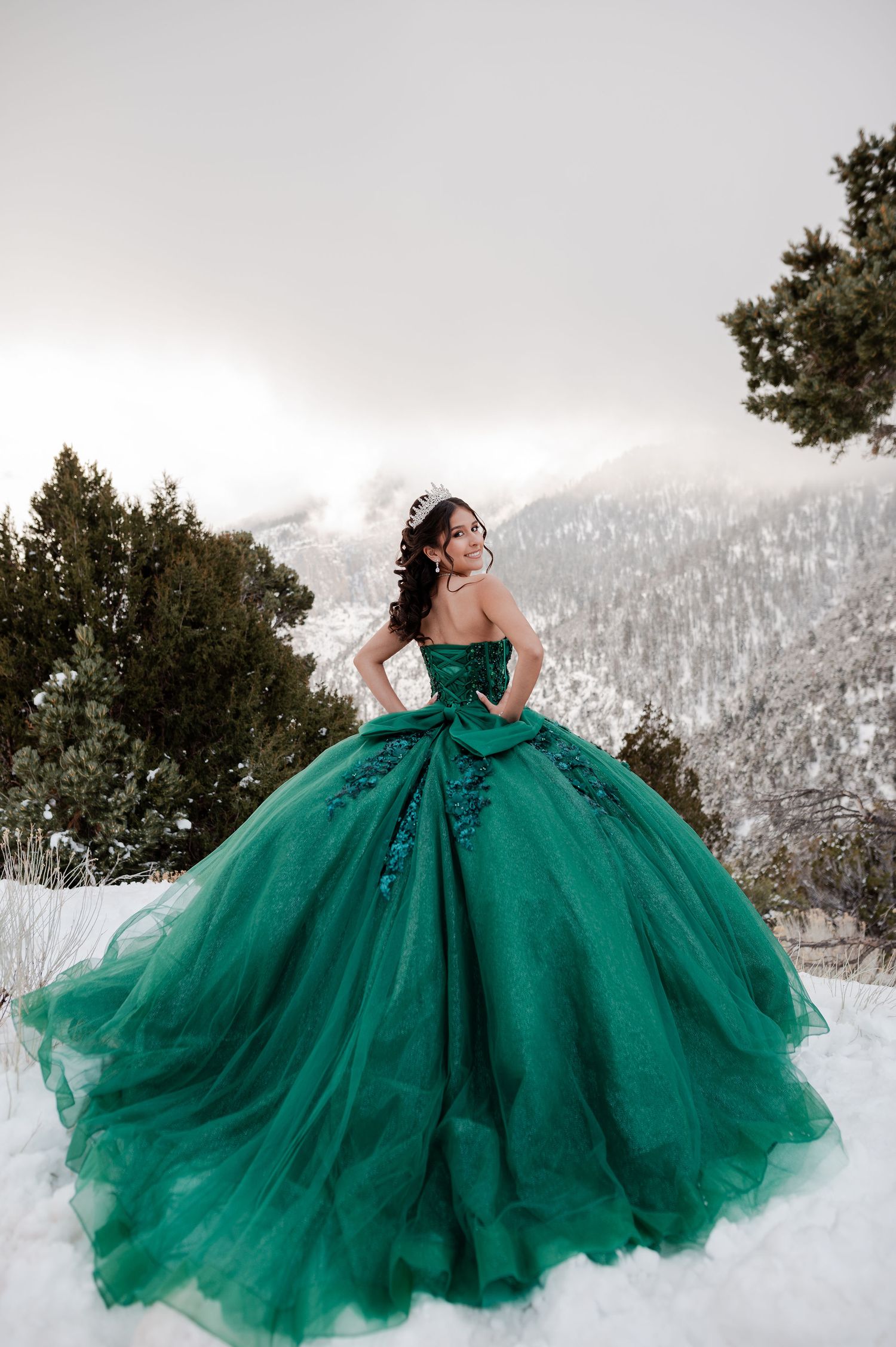 Smiling woman in emerald green strapless quinceañera gown with tiara stands in snowy mountain setting.