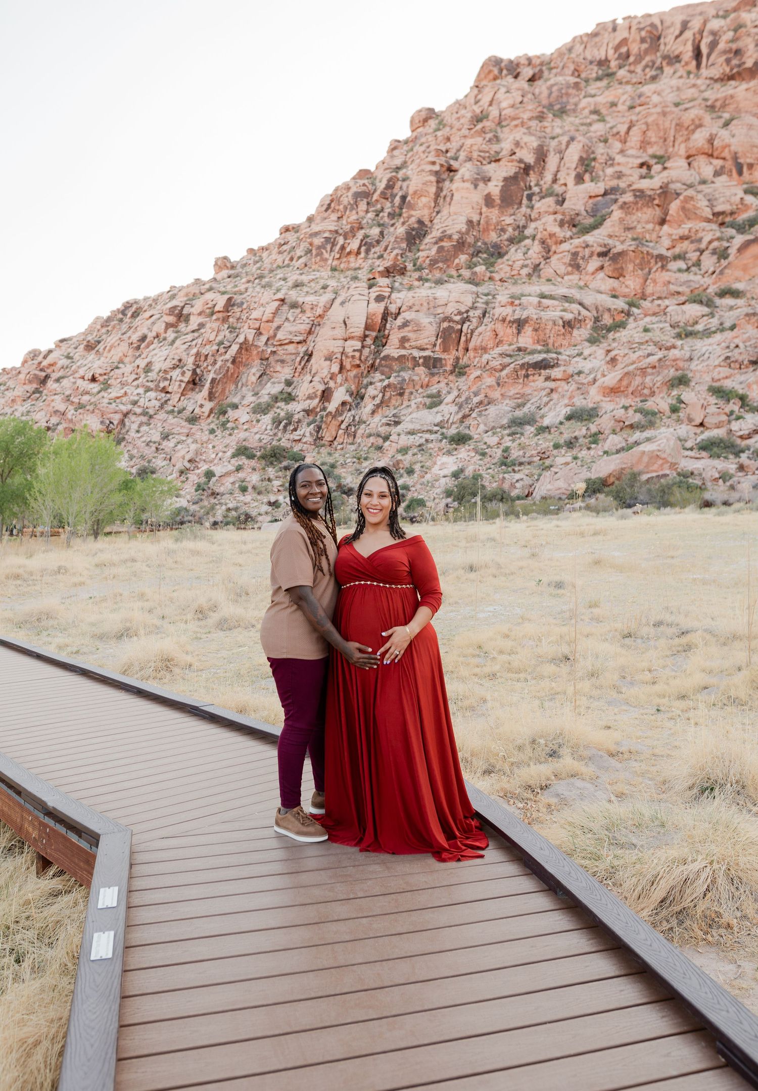 Pregnant woman in red gown with companion posing on boardwalk at Red Rock Canyon.