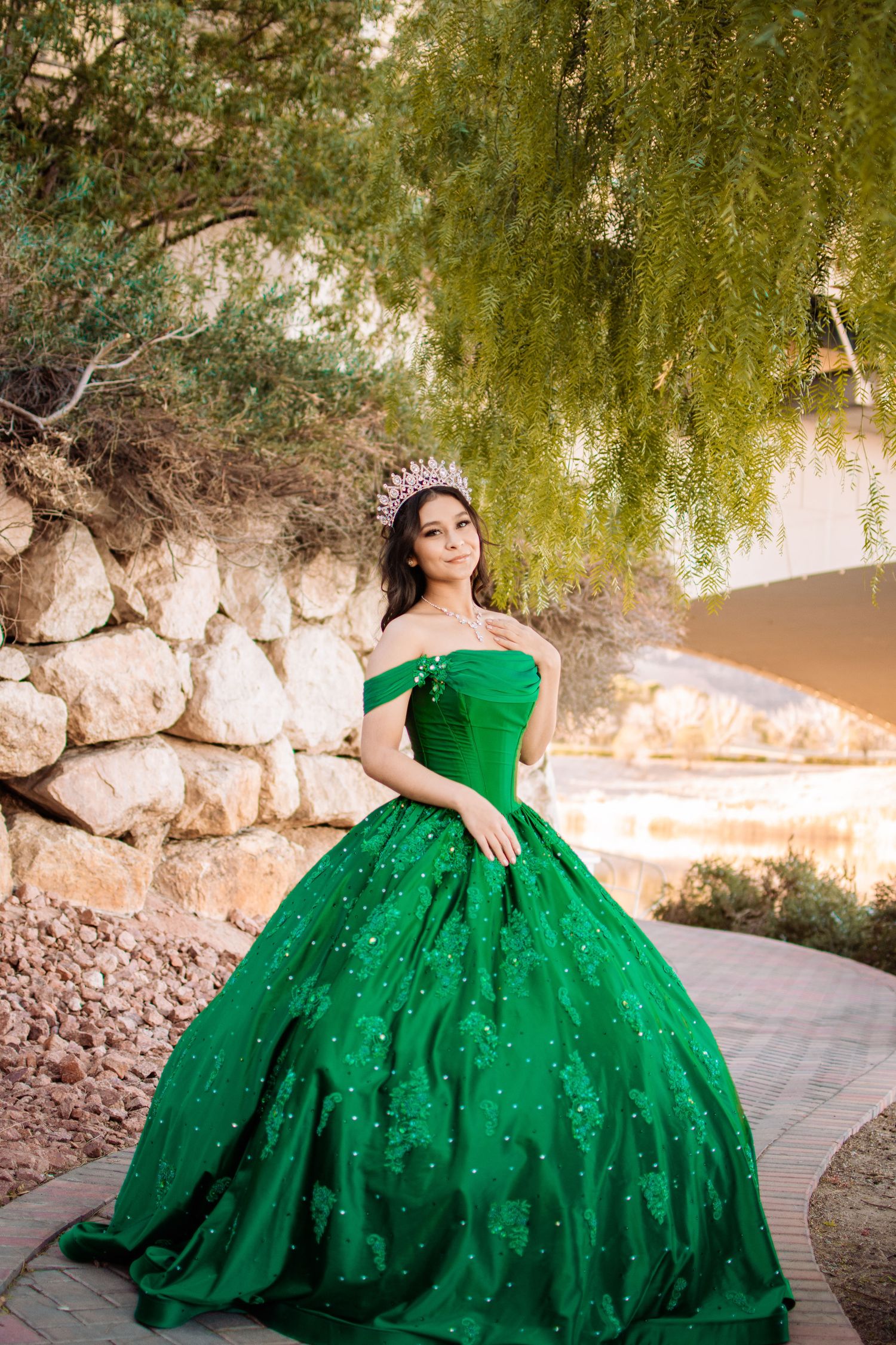 Young woman in a green quinceañera ball gown and tiara poses along a stone garden walkway.