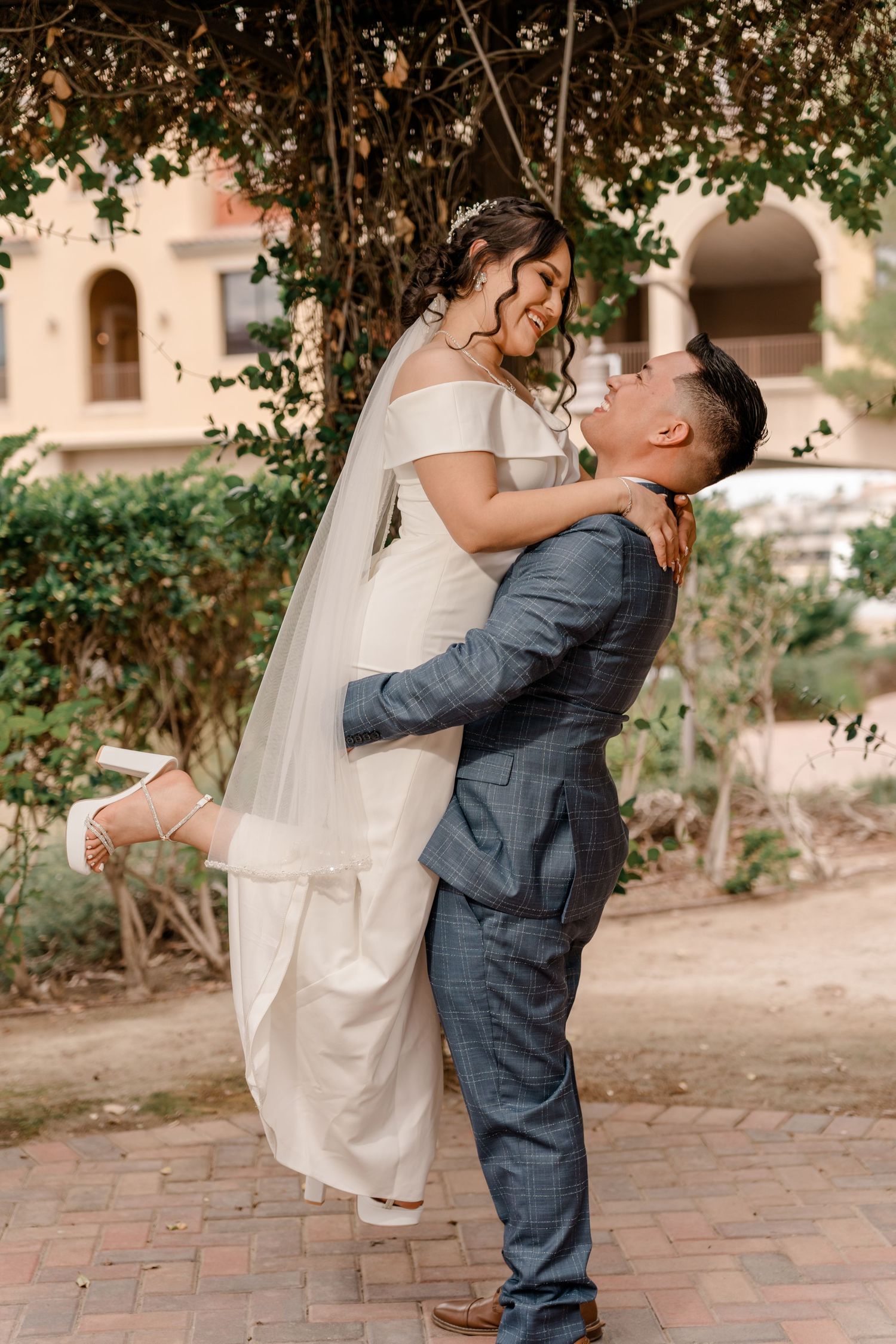 Groom lifts smiling bride in white gown and veil beneath a lush garden canopy at a Las Vegas venue.