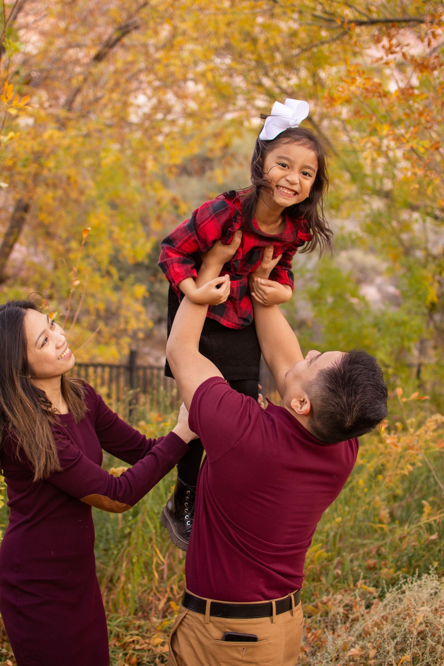 A father lifts a smiling girl in a red plaid dress while mother watches during an autumn outdoor session.
