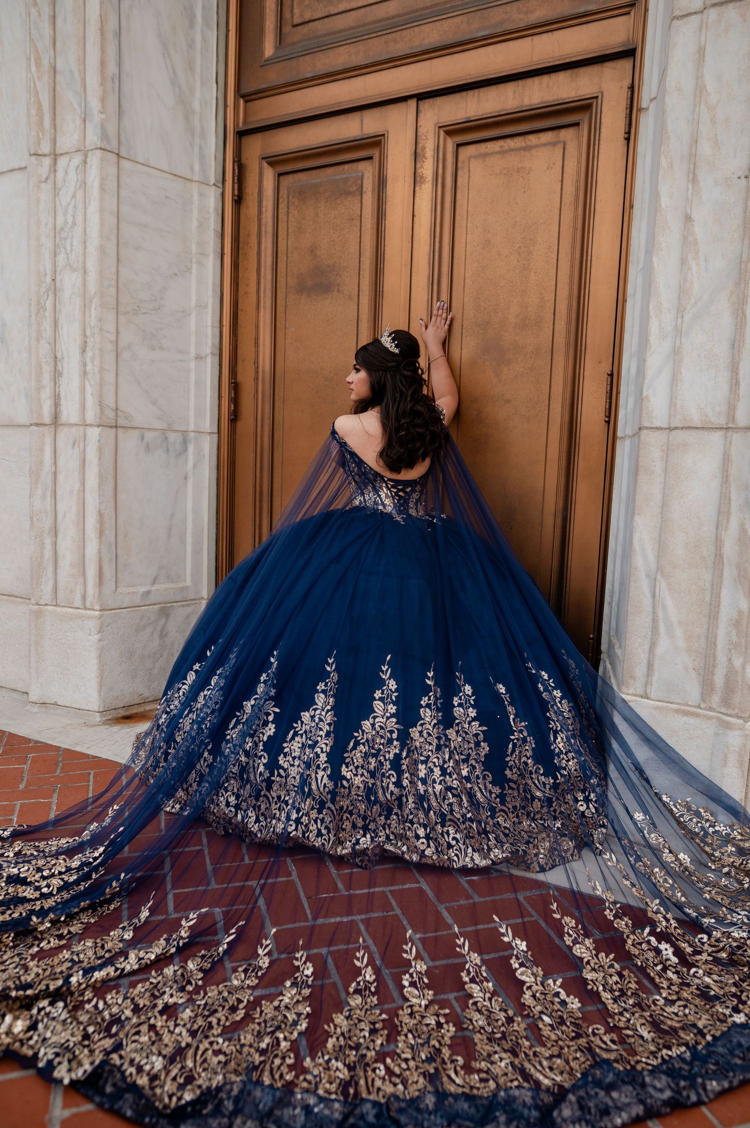 Young woman in navy blue and gold embroidered quinceañera ballgown with cape train poses at ornate wooden doors.