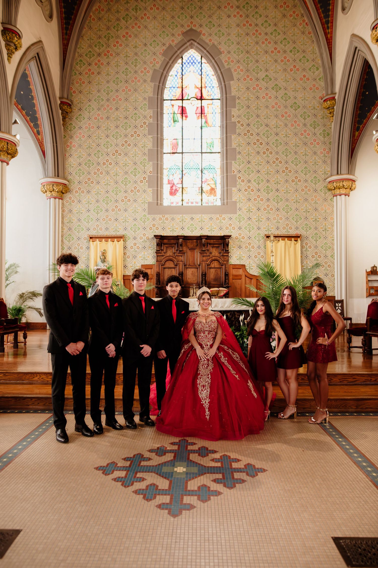 Group of teens in red and black formal attire posing inside an ornate church for a quinceañera celebration.