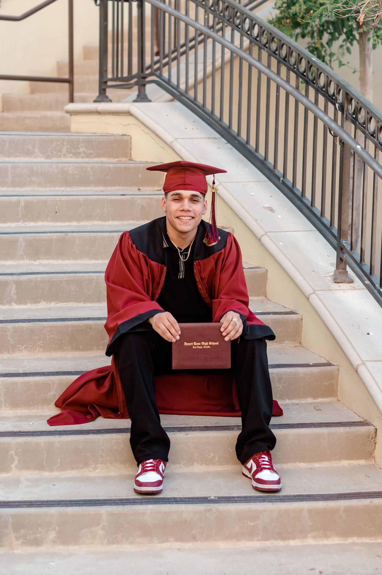 Smiling graduate in red cap and gown holds diploma while seated on stone steps.