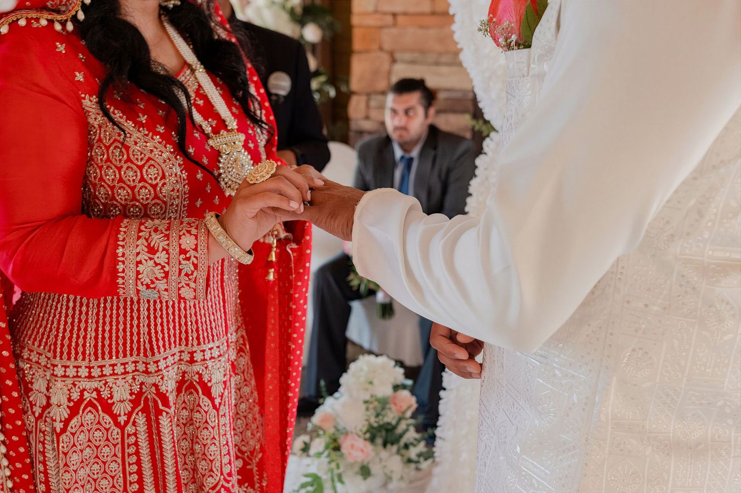 Bride in red embroidered lehenga and groom in white exchanging rings during a wedding ceremony.
