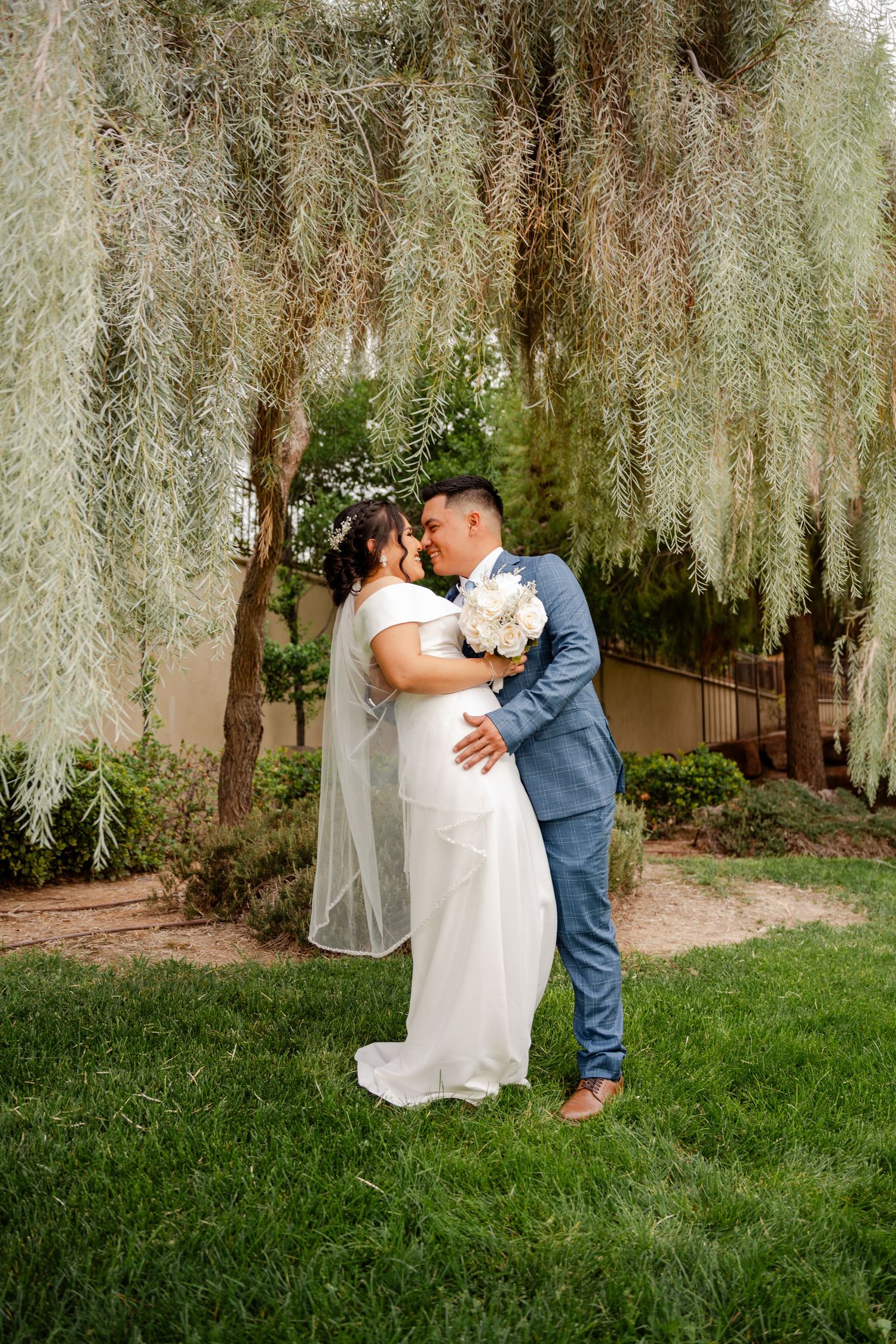 Bride and groom share a kiss under hanging willow branches on a lush green lawn.