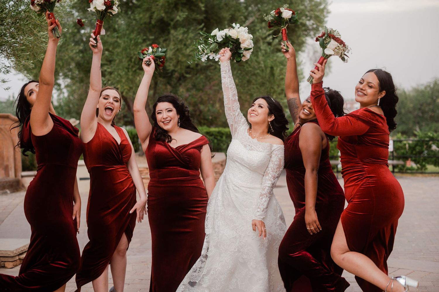 Bride and bridesmaids in burgundy velvet dresses celebrate joyfully outdoors raising bouquets.