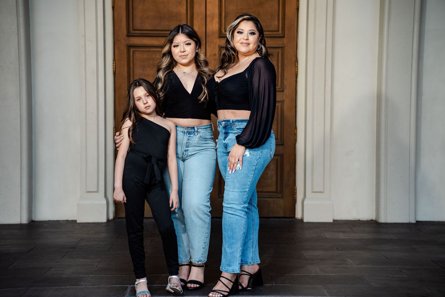 Three females pose together indoors in matching black crop tops and blue jeans near a wooden door.
