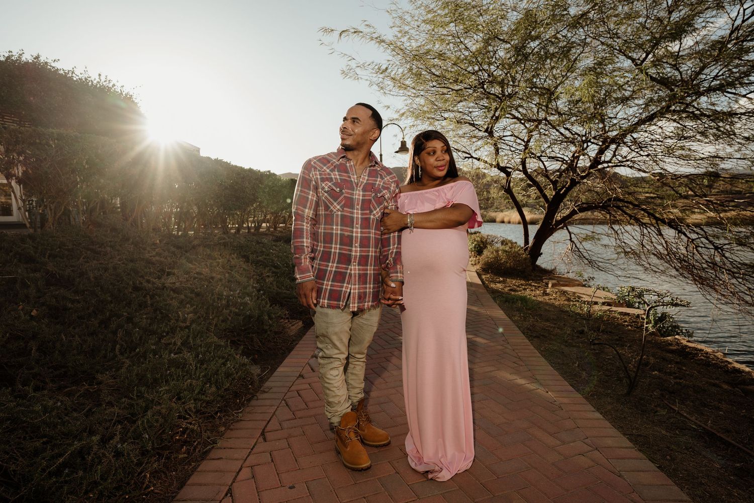 Couple posing on brick path at sunset, pregnant woman in pink gown embraced by partner near waterfront.