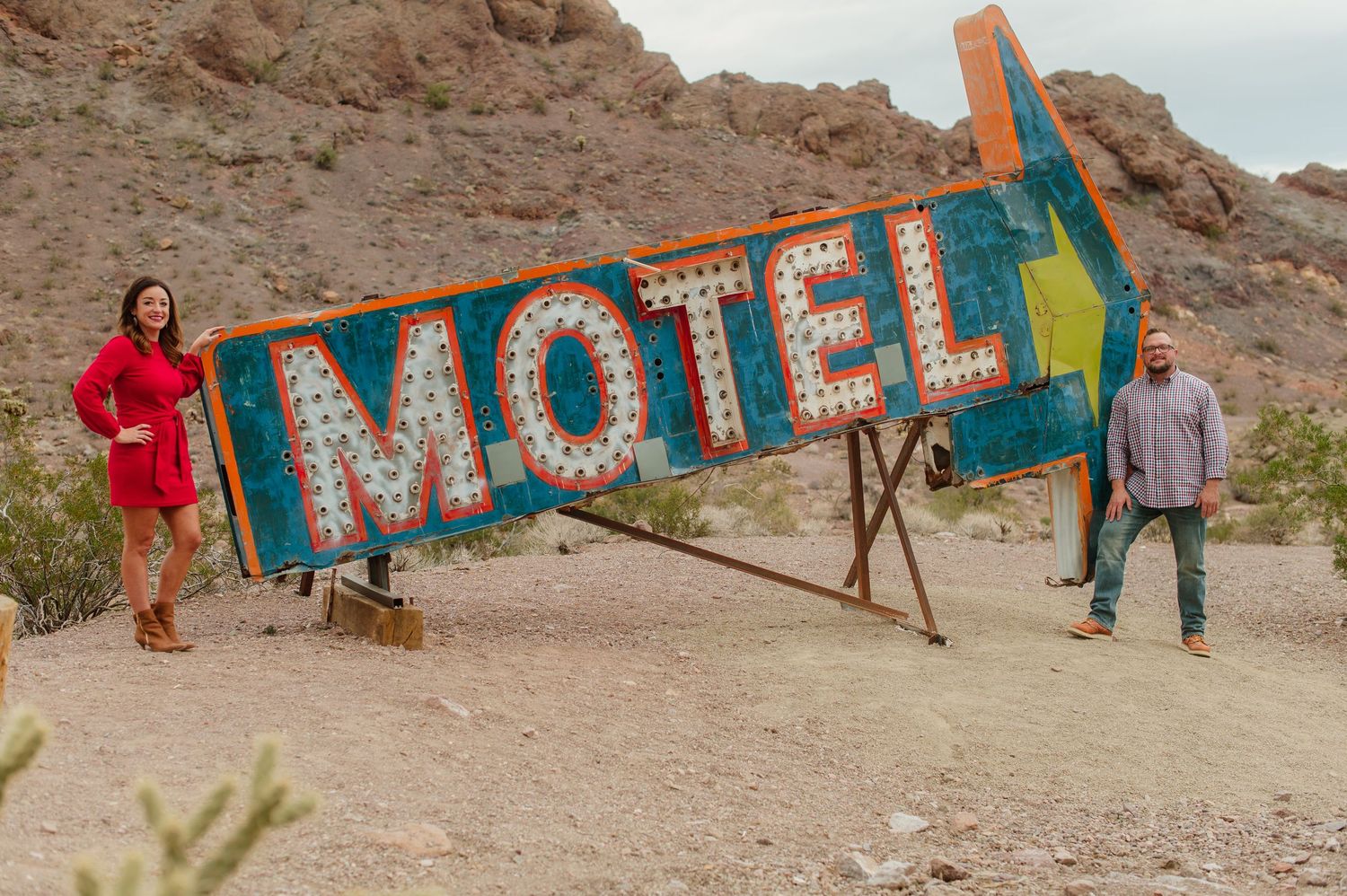 Couple posing on either side of a large vintage neon Motel sign in a rocky desert landscape.