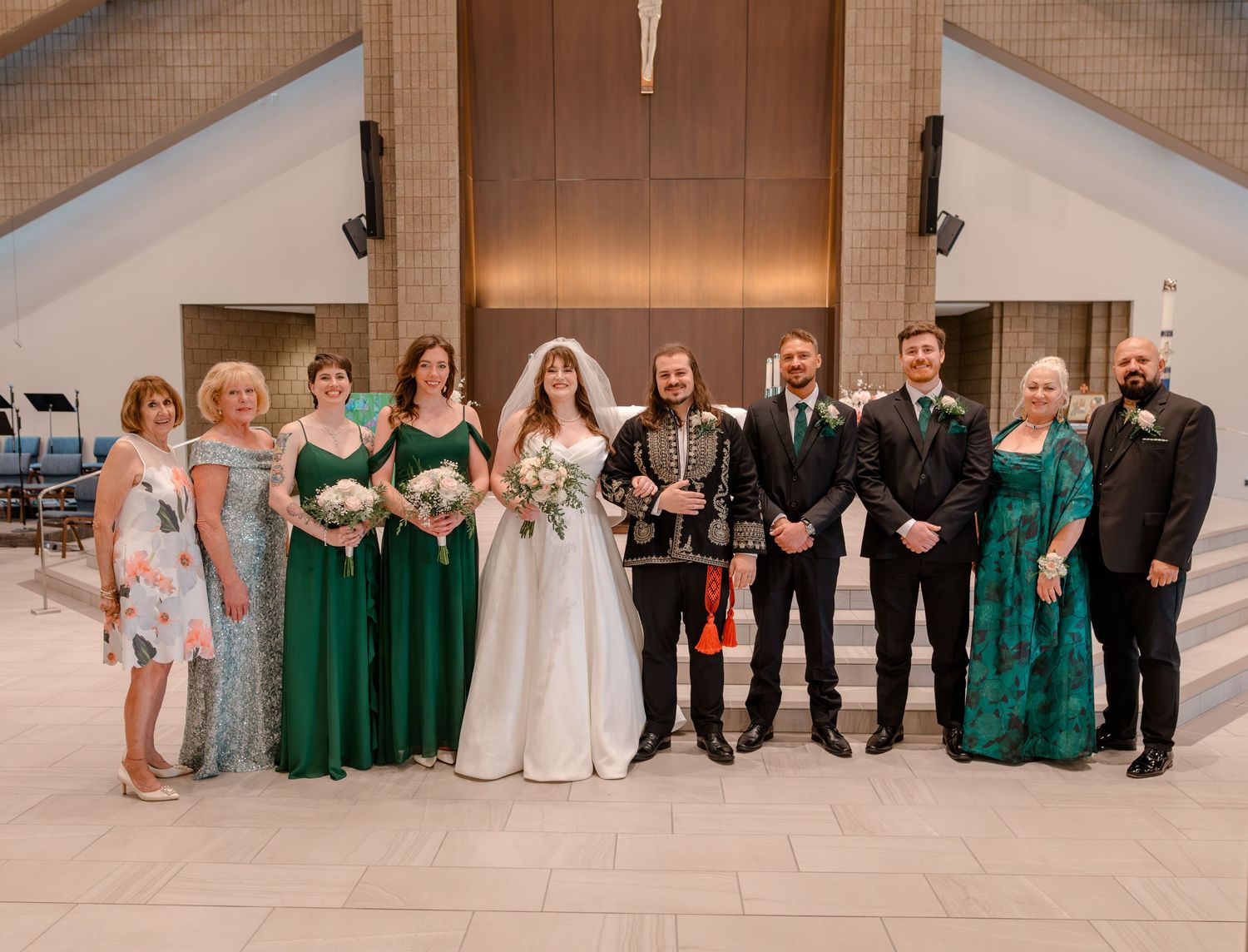 Wedding party poses in church, bride in white gown with bridesmaids in emerald green dresses and groomsmen in black suits.