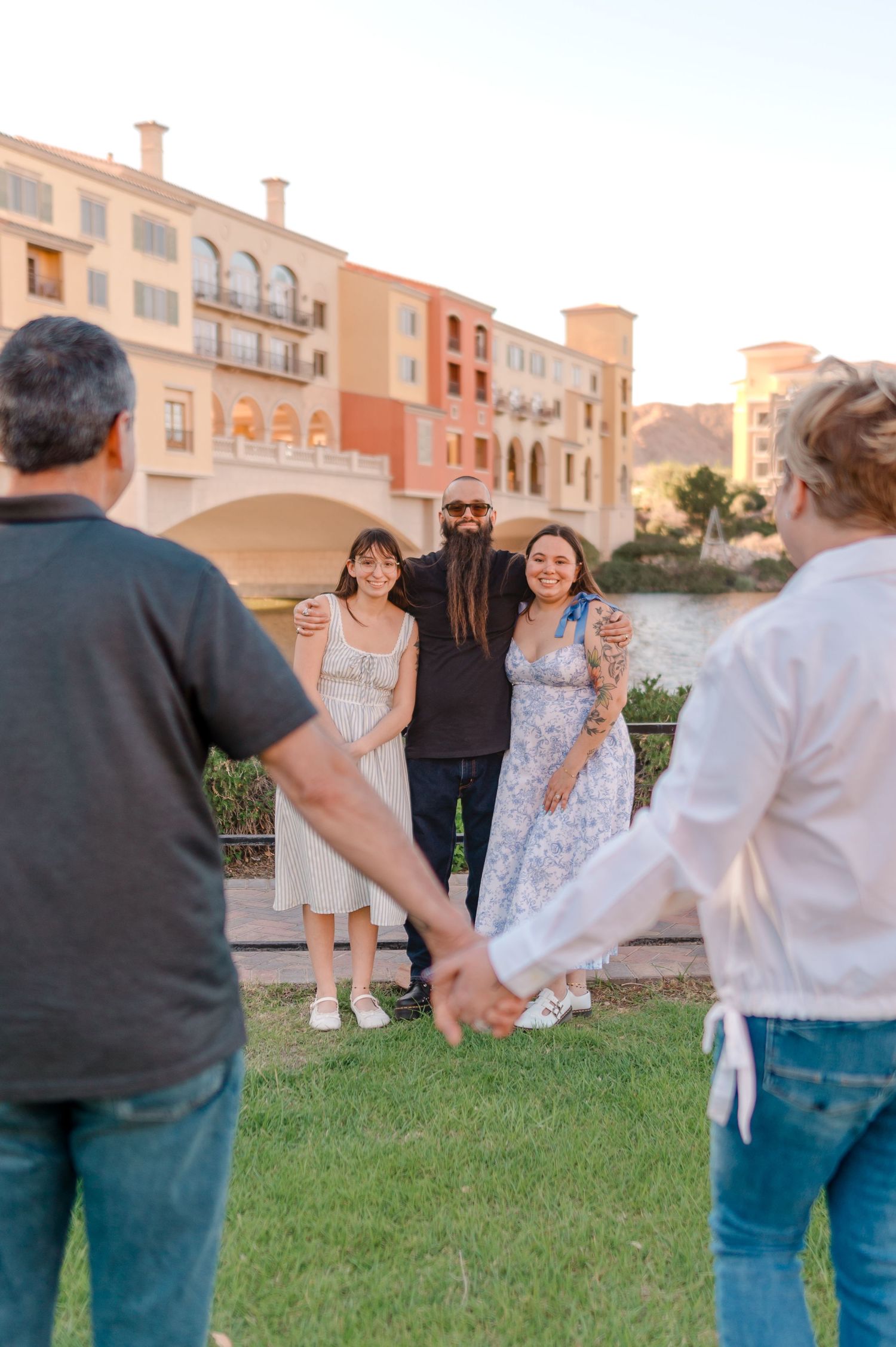 Family group poses on green lawn with Italian-style bridge and colorful buildings at golden hour.