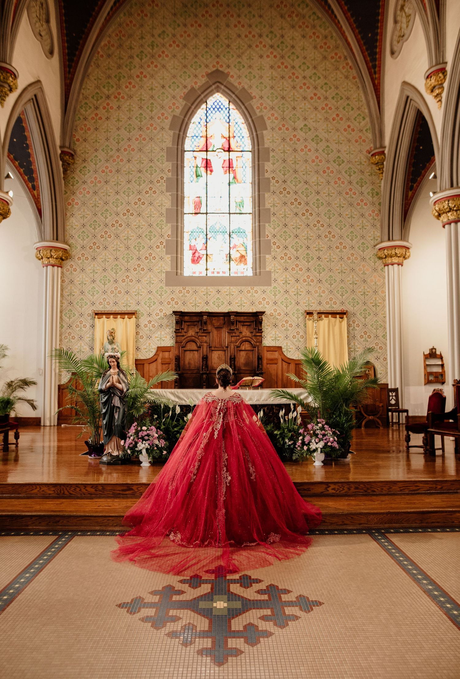 Quinceañera in red gown standing before ornate church altar with stained glass window above.