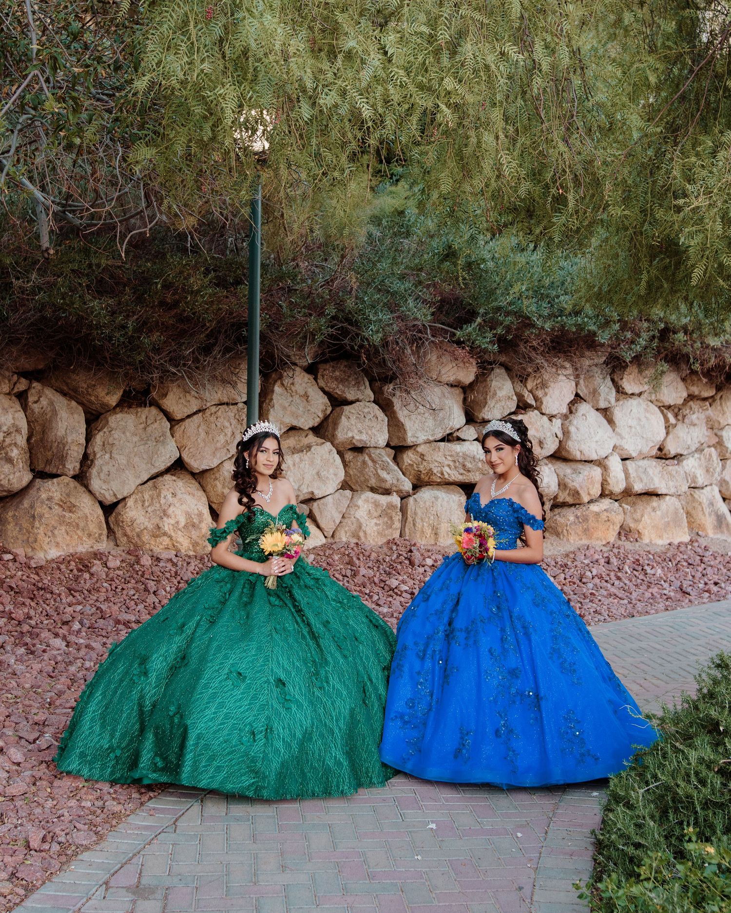 Two young women in emerald green and royal blue quinceañera ball gowns pose together outdoors with bouquets.
