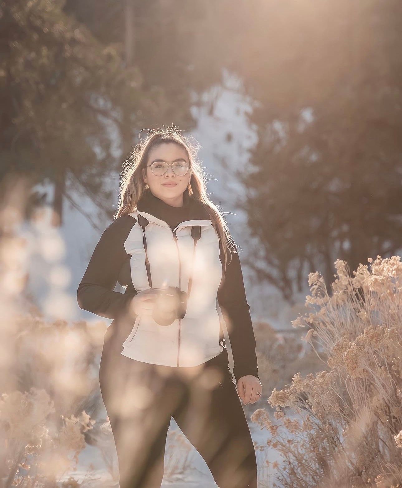 Young woman in white vest and black outfit posing in snowy winter forest with golden sunlight.