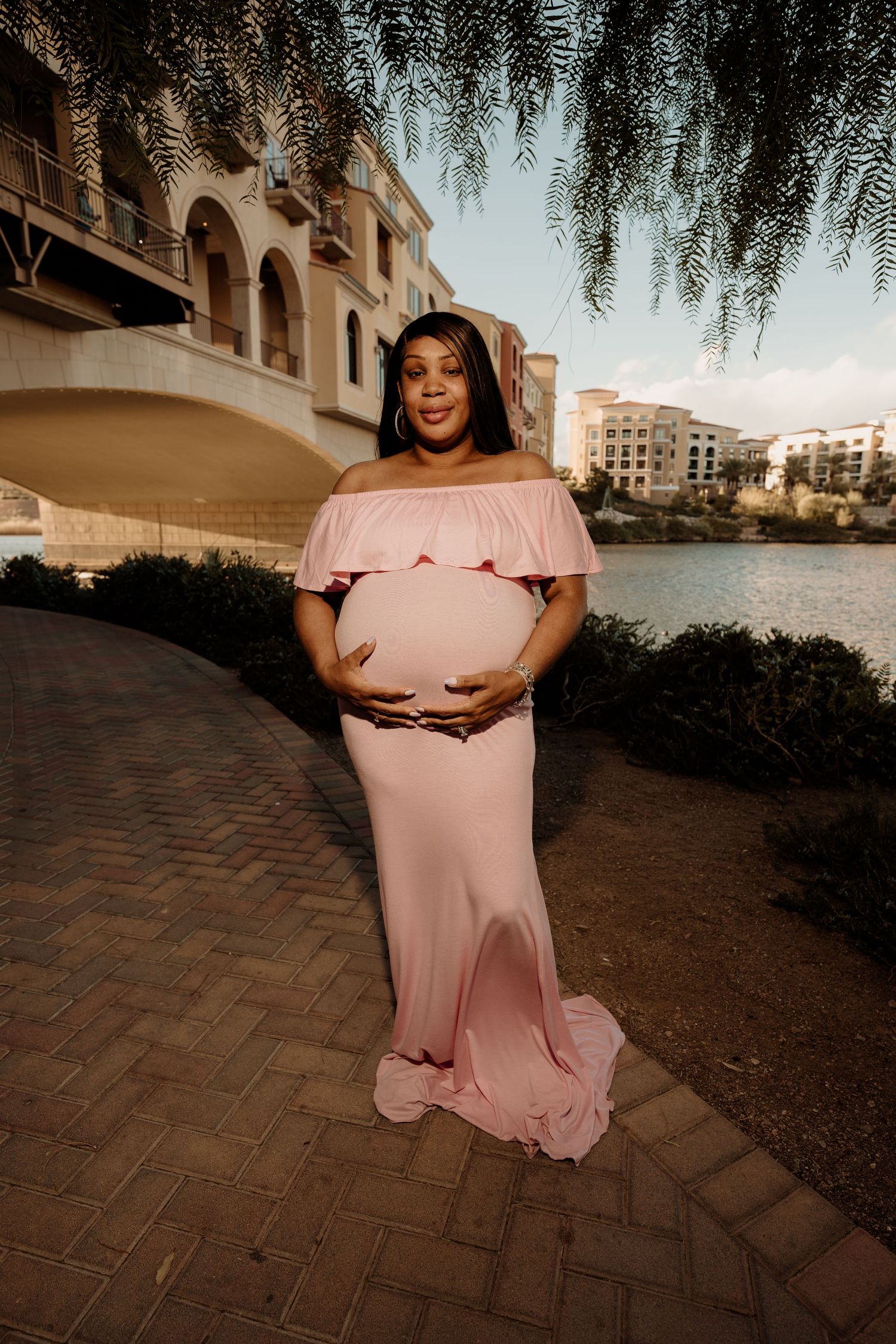 Pregnant woman in elegant pink maternity dress cradling bump on brick path near waterfront buildings at dusk.