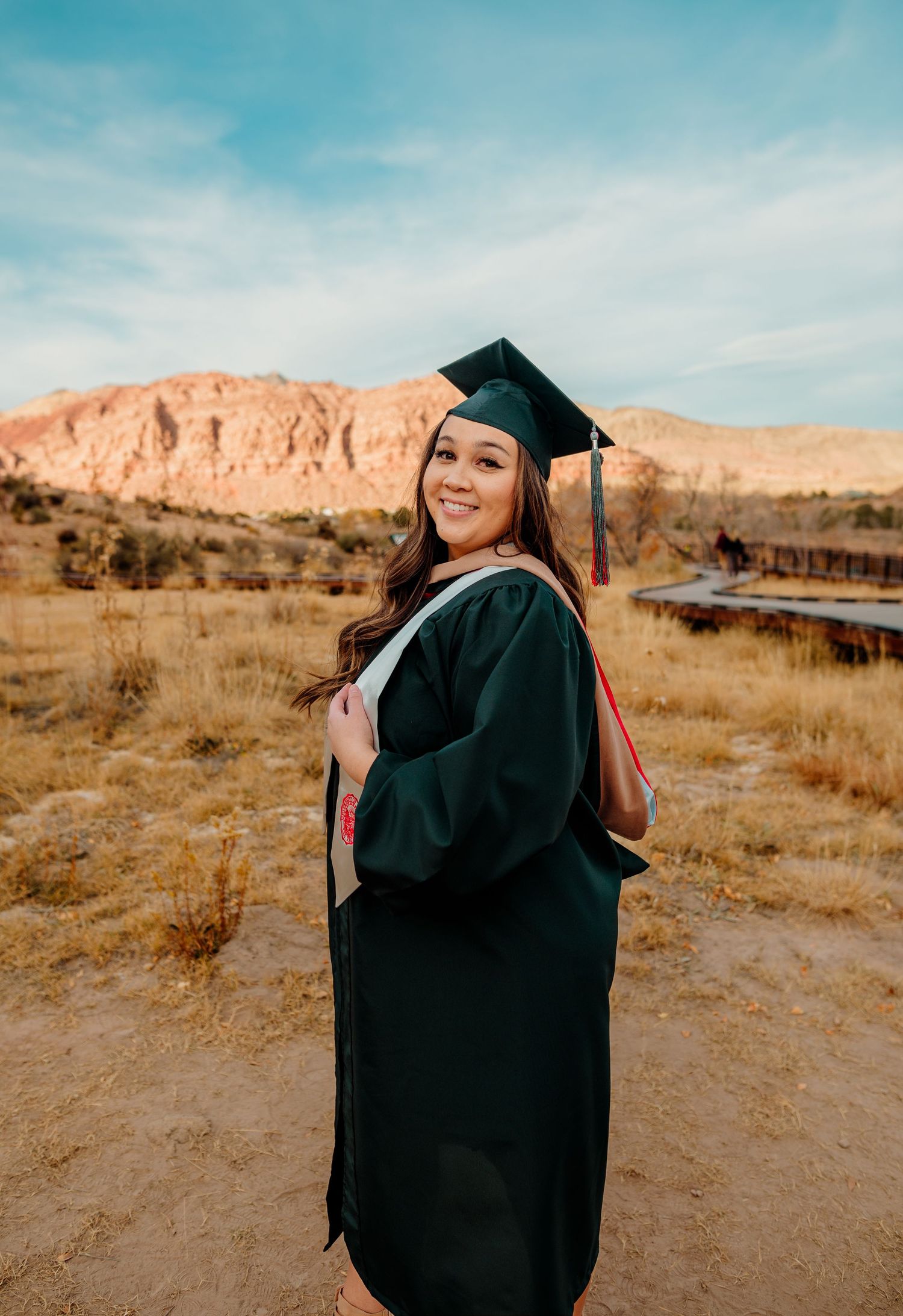 Smiling female graduate in dark green cap and gown poses at Red Rock Canyon landscape.