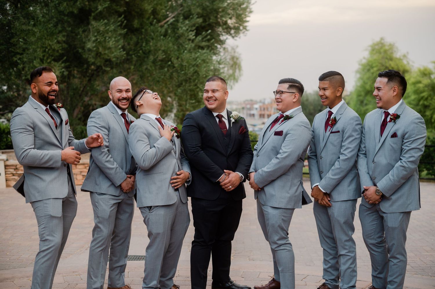 Groom in black suit stands with groomsmen in matching gray suits laughing outdoors.
