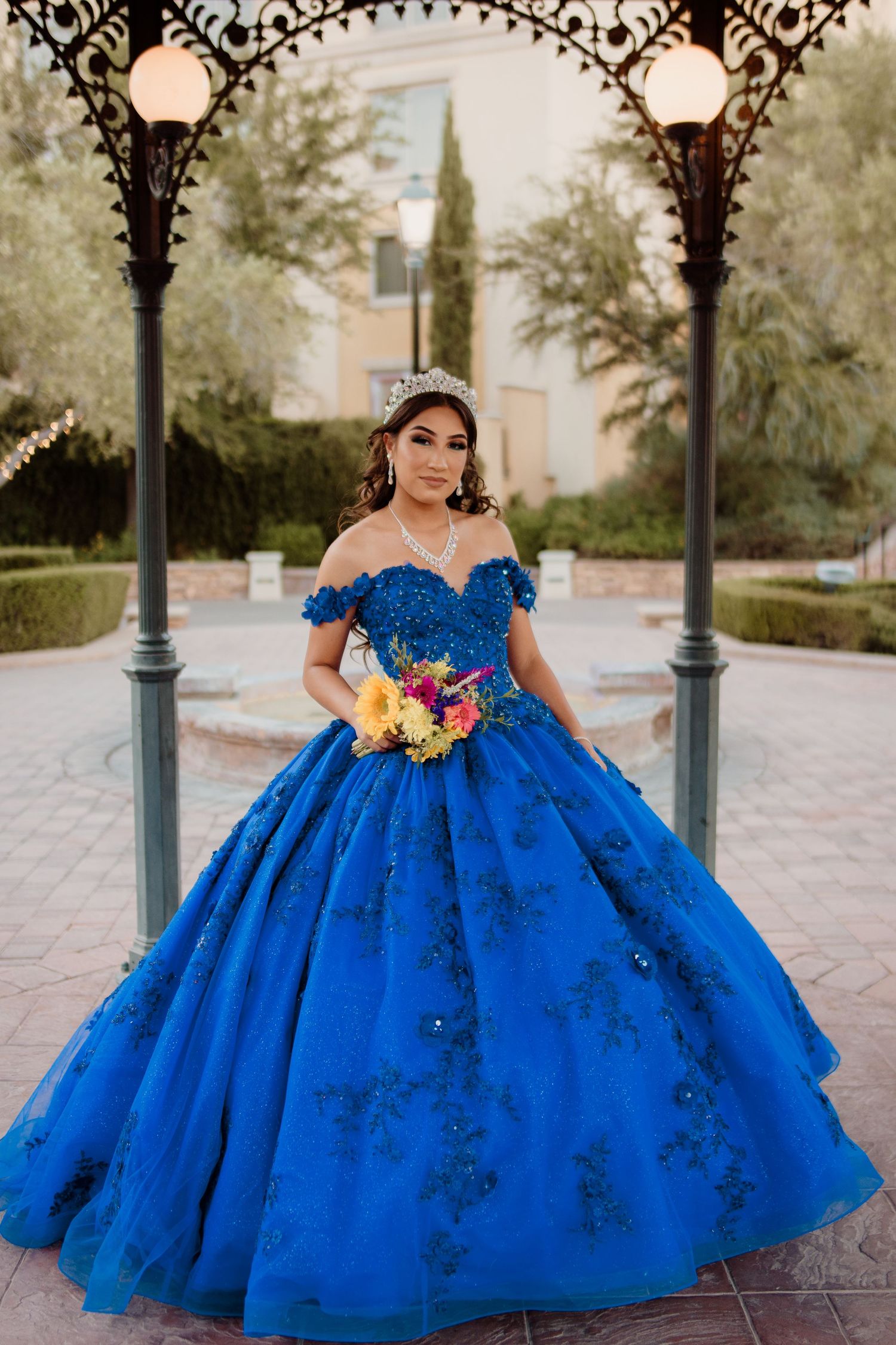 Young woman in royal blue off-shoulder quinceañera ball gown holds colorful bouquet under ornate iron archway.