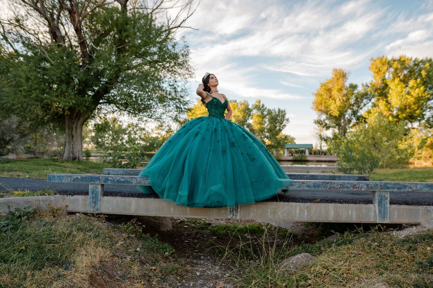 Young woman in voluminous emerald green quinceañera gown poses on a wooden bridge outdoors.