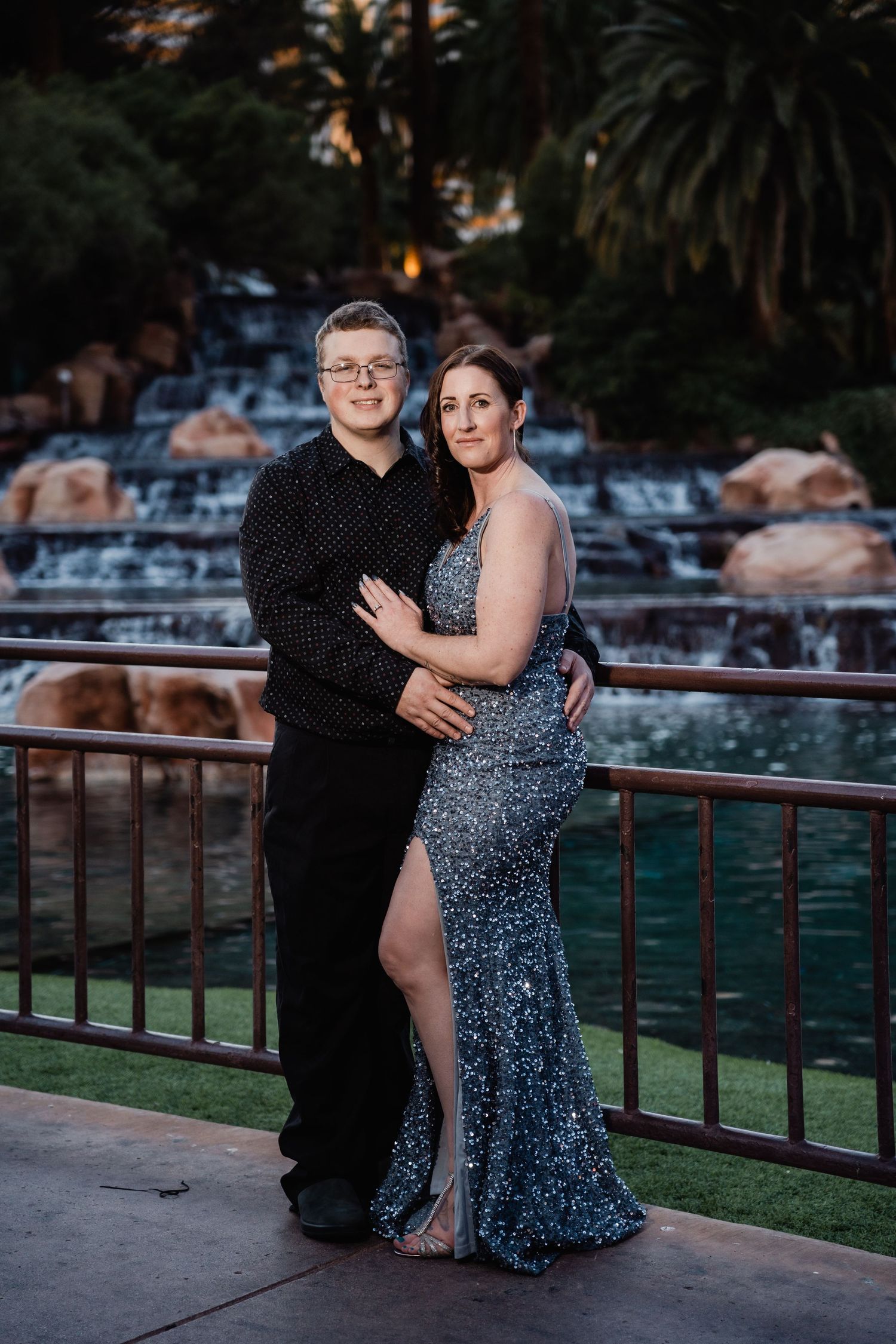 Couple posing elegantly in formal wear before a cascading waterfall backdrop at dusk.
