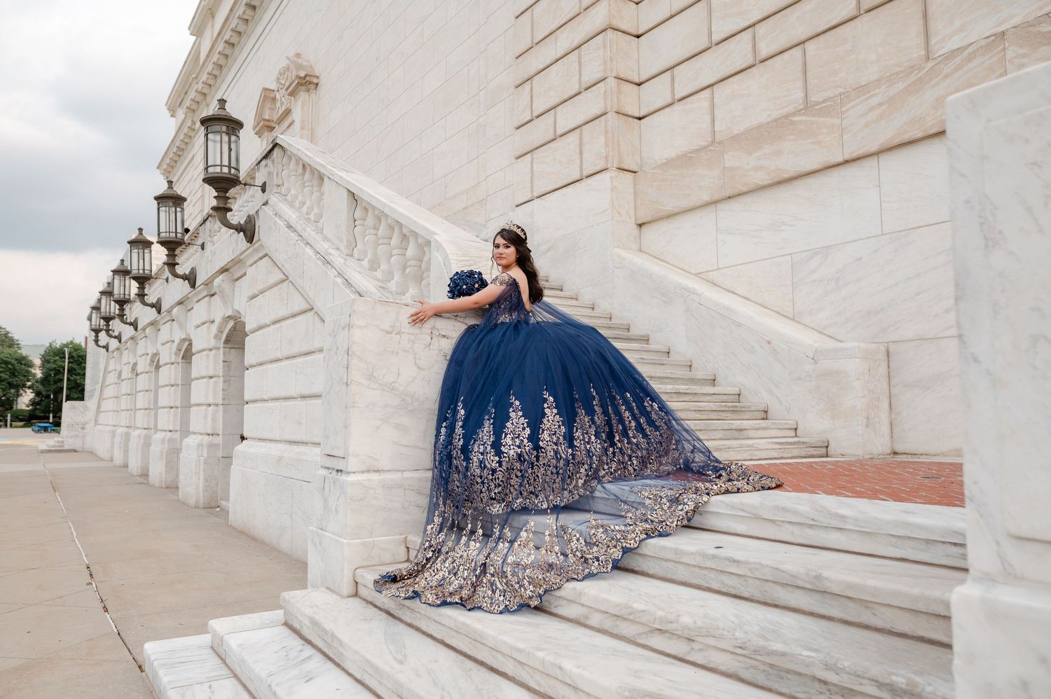 Young woman in navy blue quinceañera ball gown with gold embroidery poses gracefully on marble steps of elegant building.