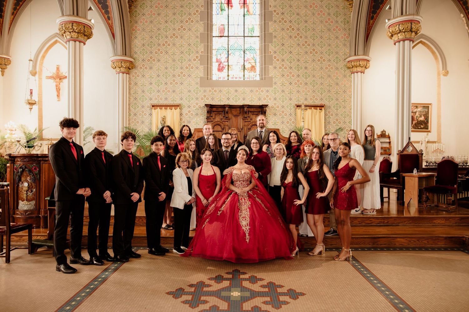 Large group photo of quinceañera court in red and black formal attire inside a beautifully decorated church.
