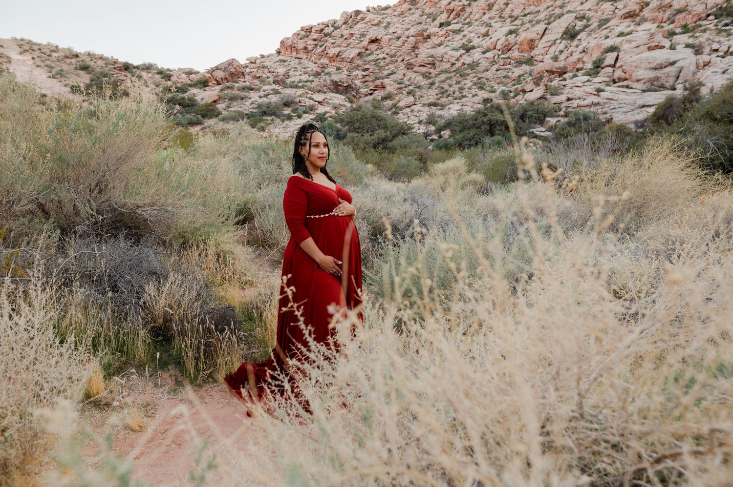 Pregnant woman in red maternity gown poses on desert trail surrounded by shrubs and red rock formations.