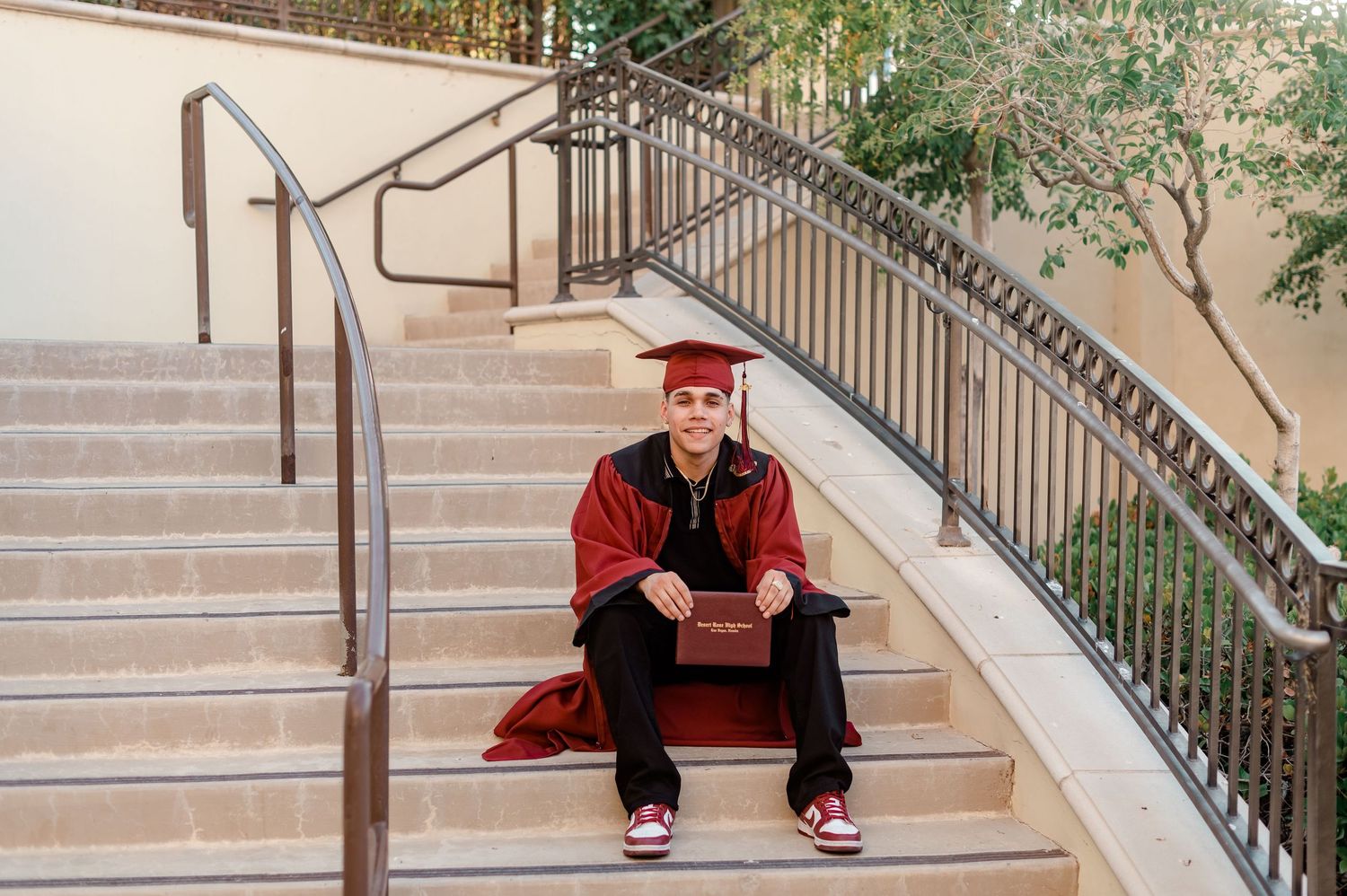 Graduate in red cap and gown sitting on outdoor steps holding diploma.