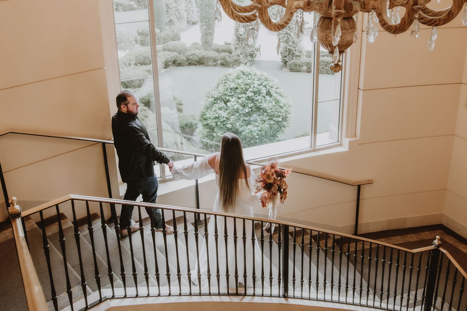 Bride and groom walk along curved staircase railing near large window with chandelier above.