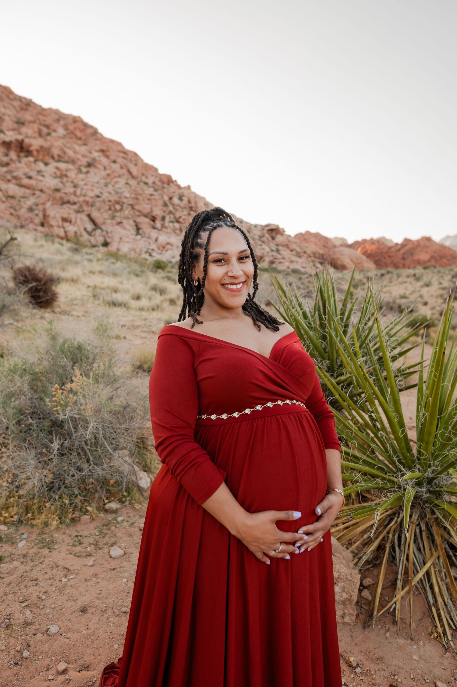 Smiling pregnant woman in elegant red maternity gown cradling baby bump at Red Rock Canyon desert.