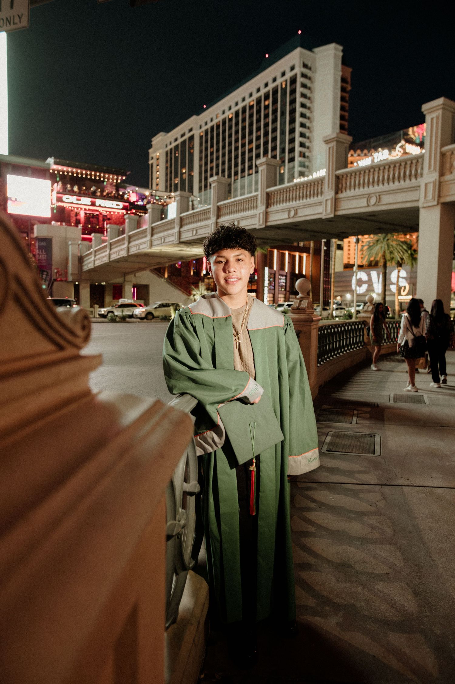 Smiling graduate in green gown poses on Las Vegas Strip sidewalk at night.