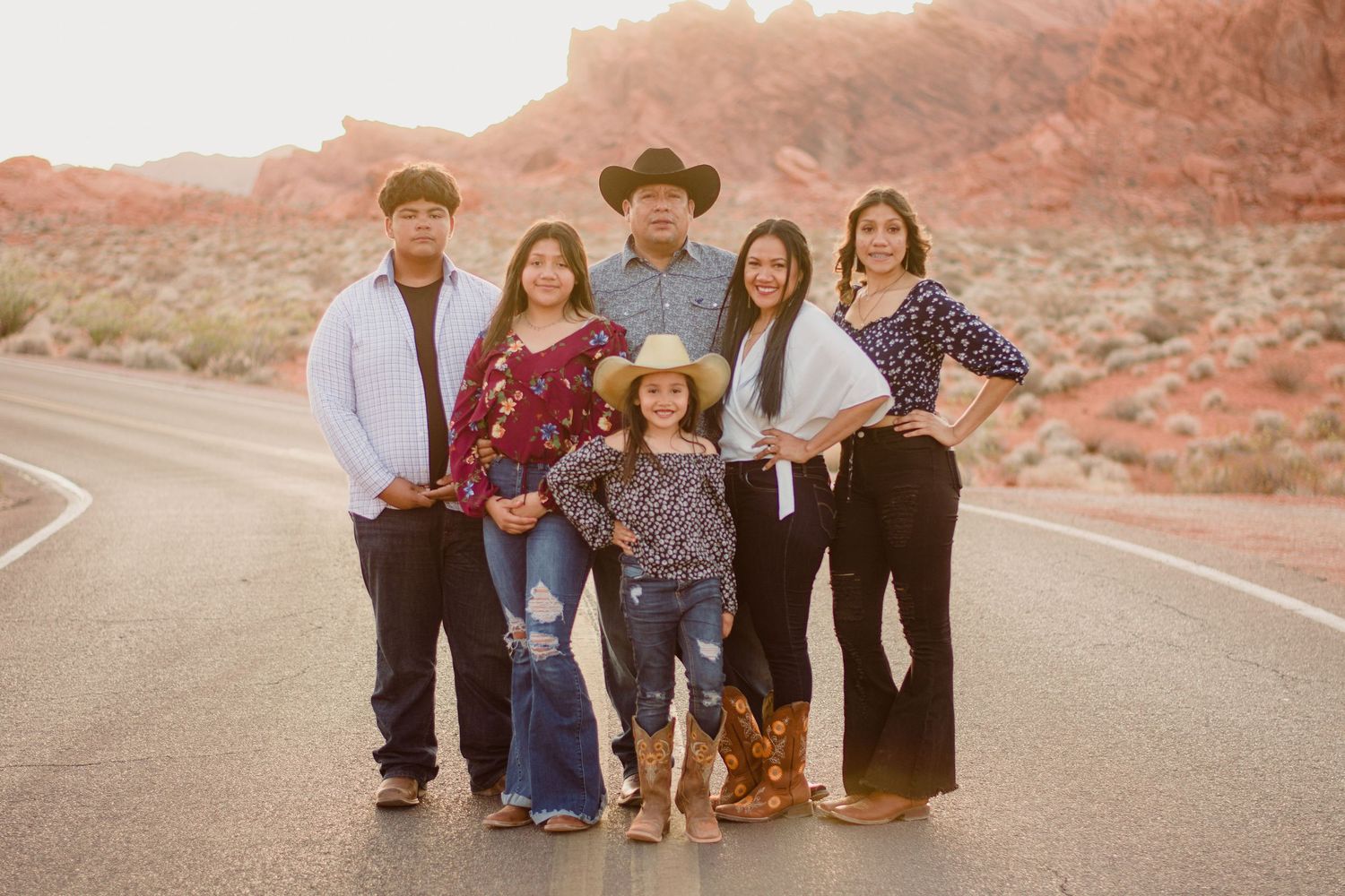 A family of six poses together on a desert road with red rock formations at golden sunset.