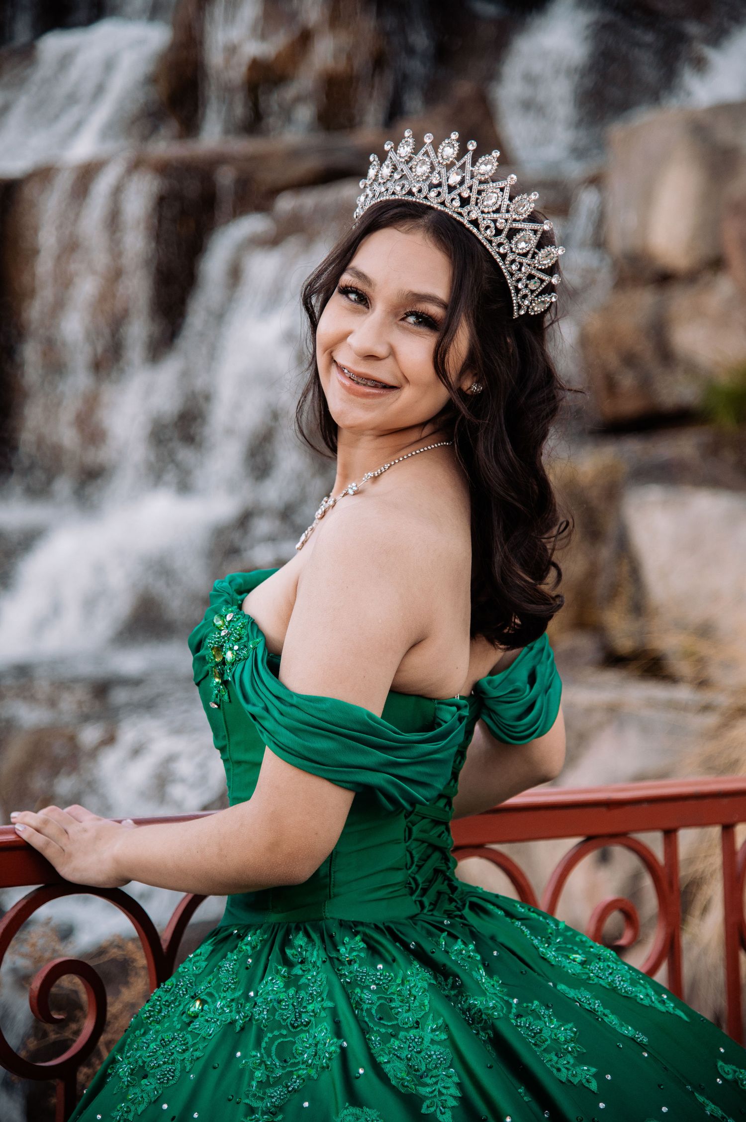 Smiling young woman in green off-shoulder quinceañera gown and crystal tiara near waterfall backdrop.