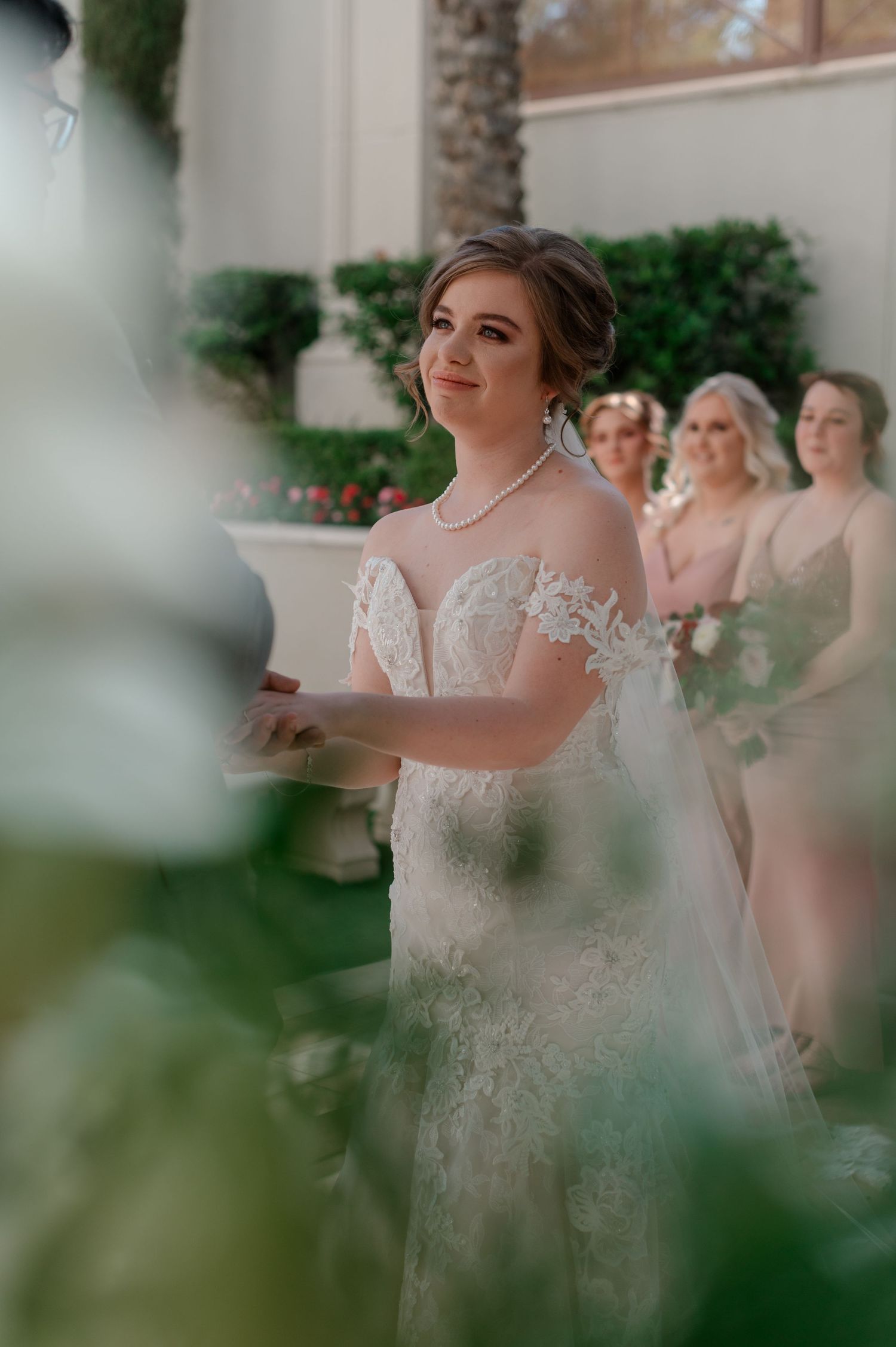 Bride in lace off-shoulder gown smiles during outdoor wedding ceremony with bridesmaids behind.