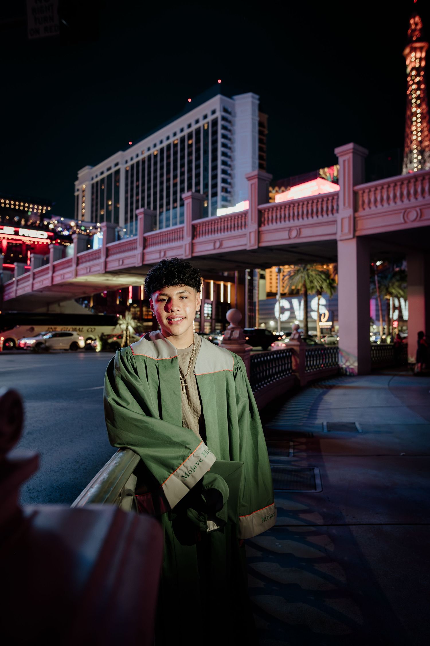 Young man in green graduation gown leans on railing along the Las Vegas Strip at night.