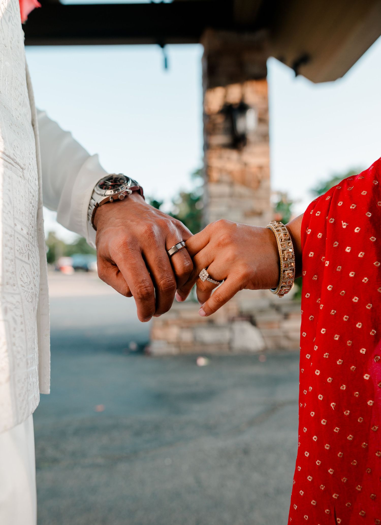A couple holds hands tenderly, wearing rings, a watch, and a gold bracelet, dressed in white and red attire.
