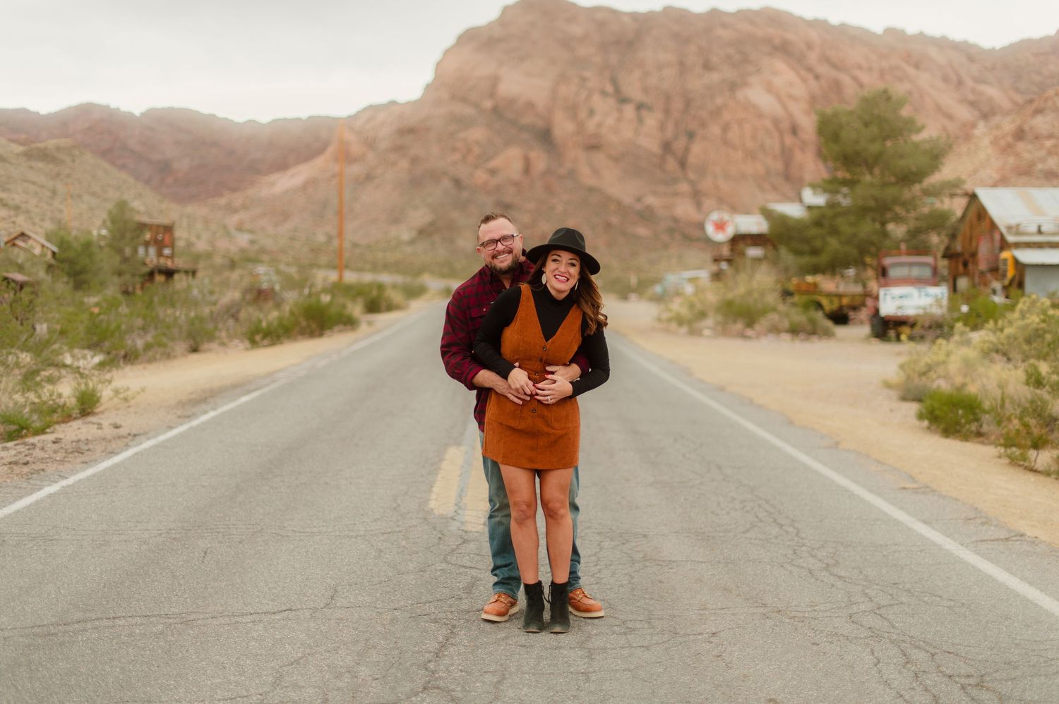 Man hugging woman in orange dress on a desert road with mountains behind them.