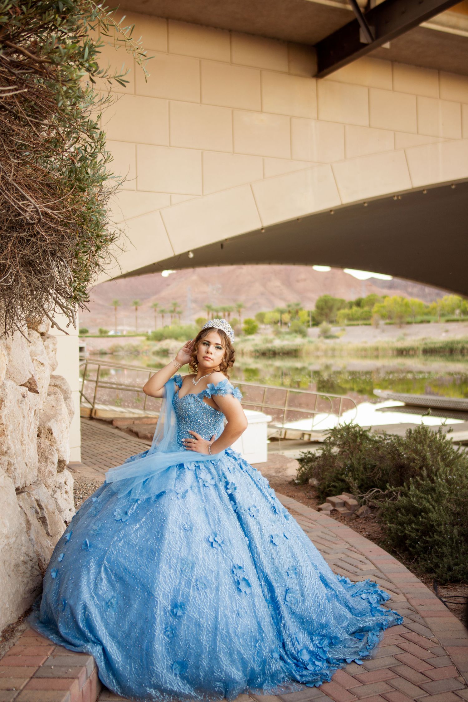 Young woman in blue off-shoulder quinceañera gown poses gracefully under modern bridge outdoors.