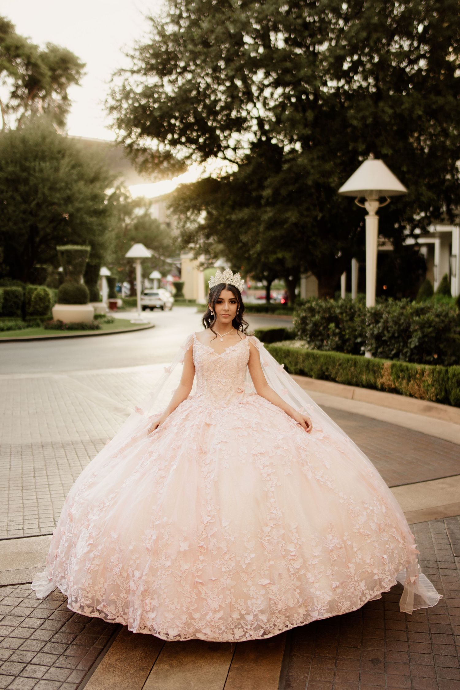 Young woman in a blush pink quinceañera ball gown with lace details and cape poses on an outdoor walkway at dusk.