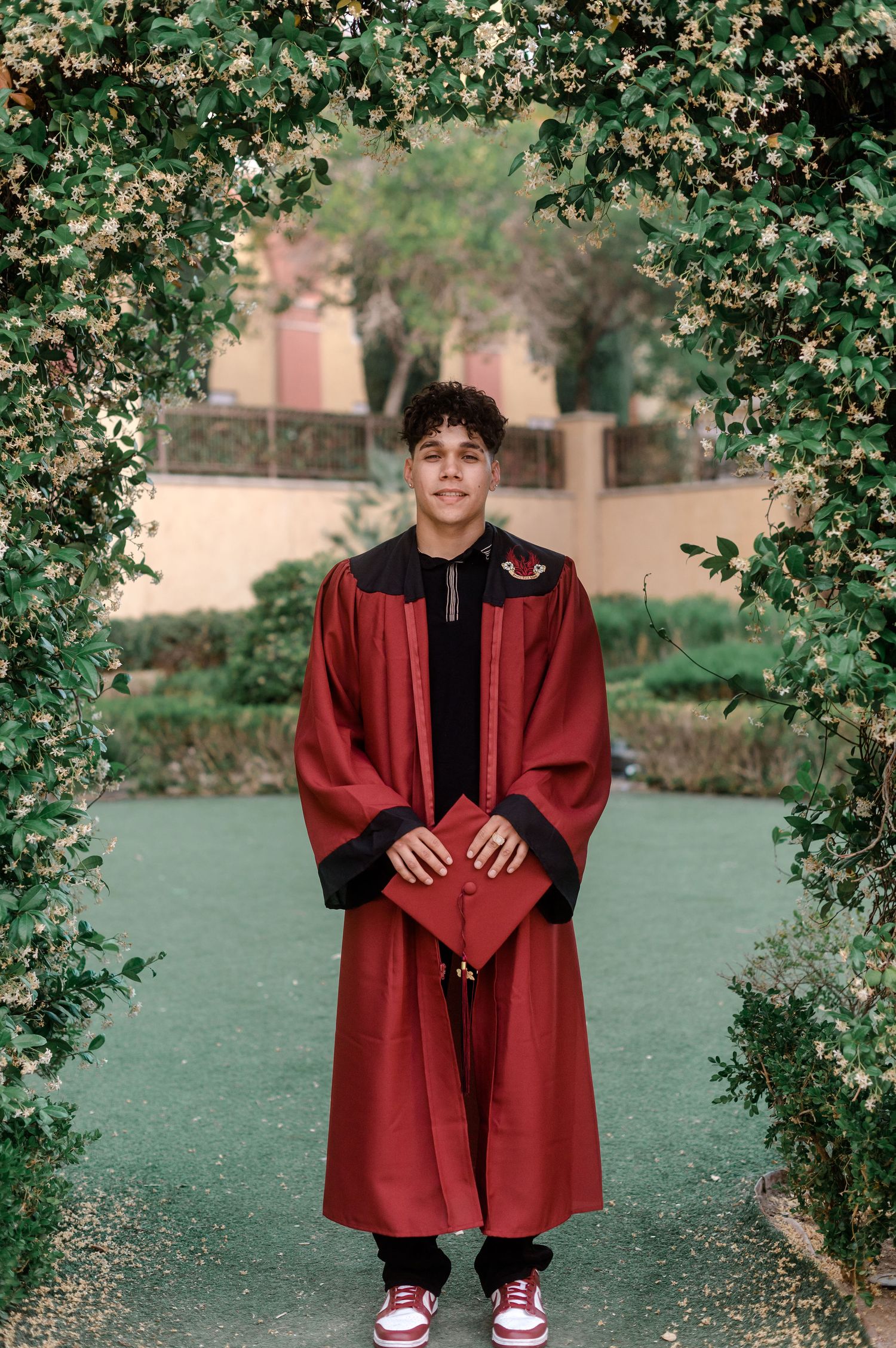 Smiling graduate in red gown holds cap under floral arch in lush garden setting.