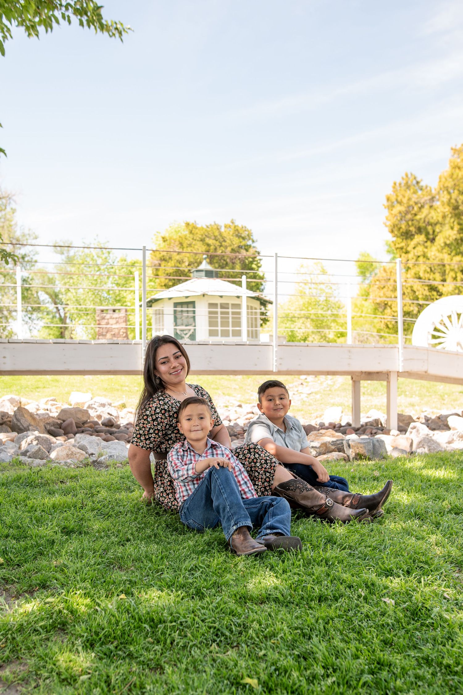 A mother sits on green grass with her two young sons at a sunny park with a gazebo backdrop.