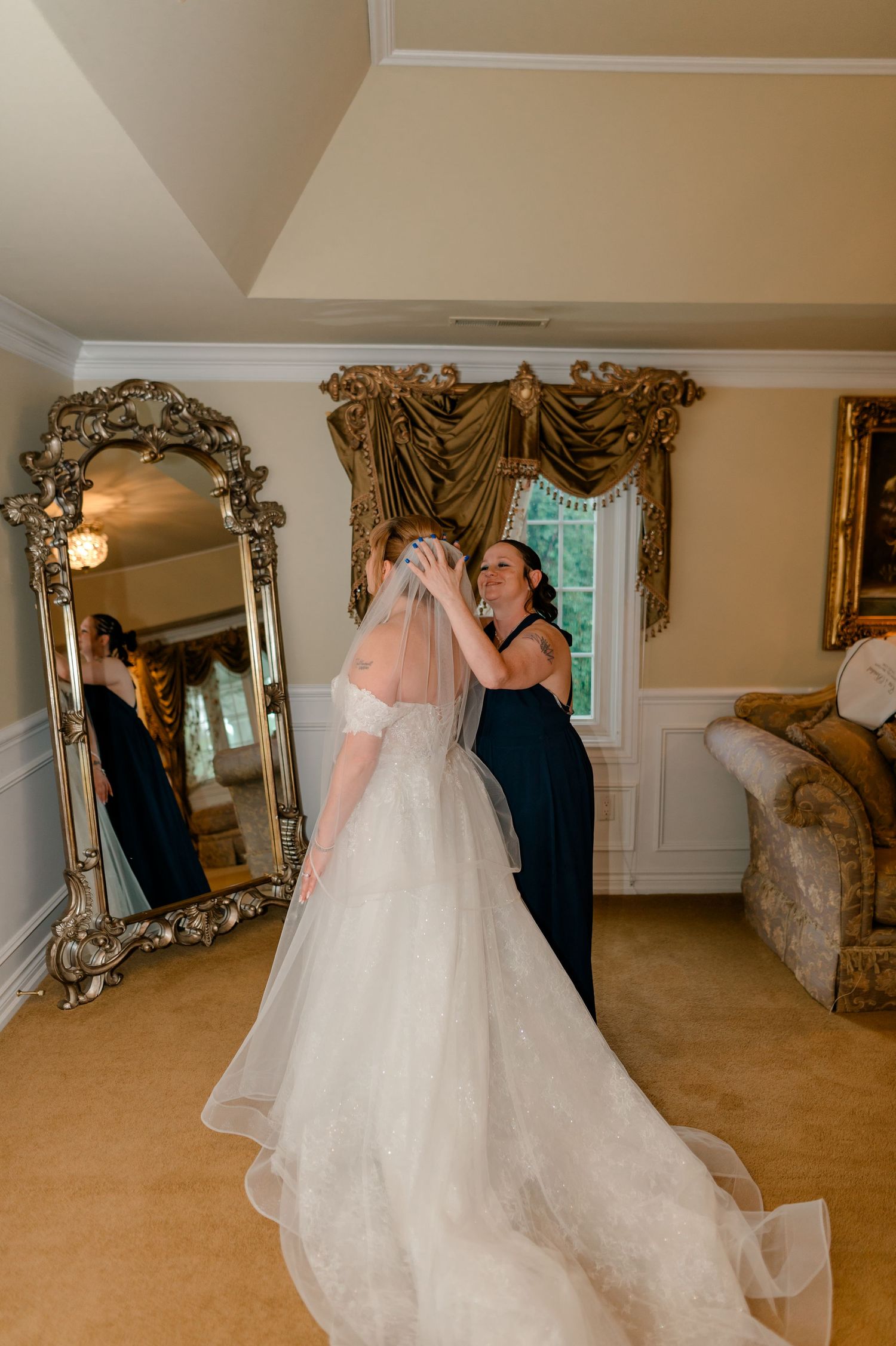 Bridesmaid helping bride with veil in elegant bridal suite with ornate gold mirror and classic decor.