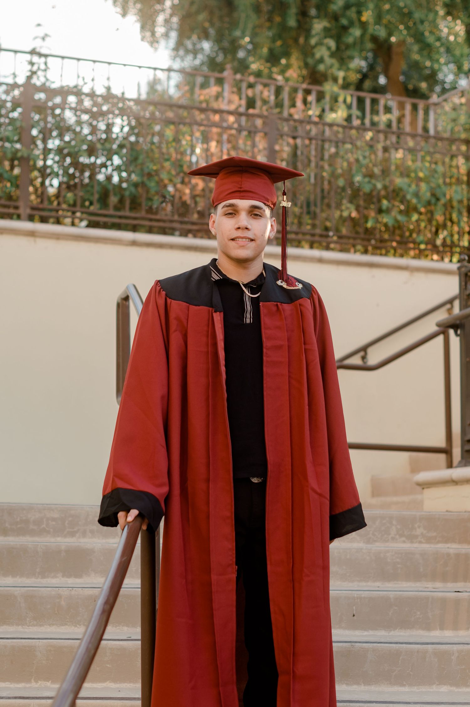 Smiling graduate in red cap and gown posing on outdoor staircase.