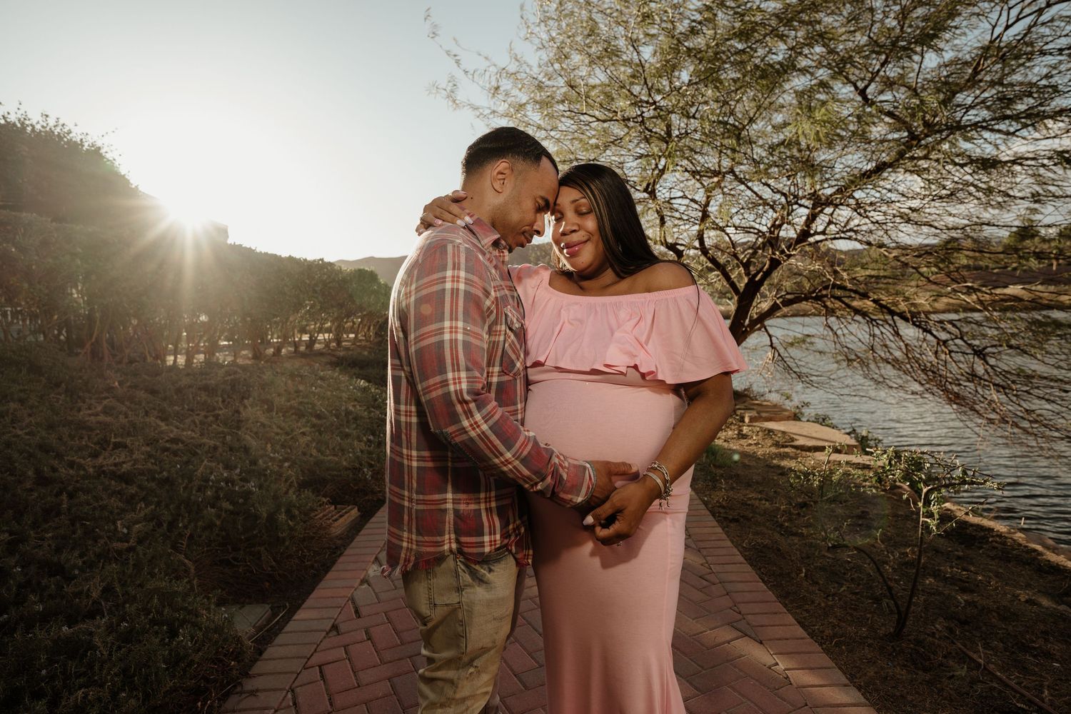 Couple sharing tender moment on brick path at sunset, man embracing pregnant woman in pink maternity gown.