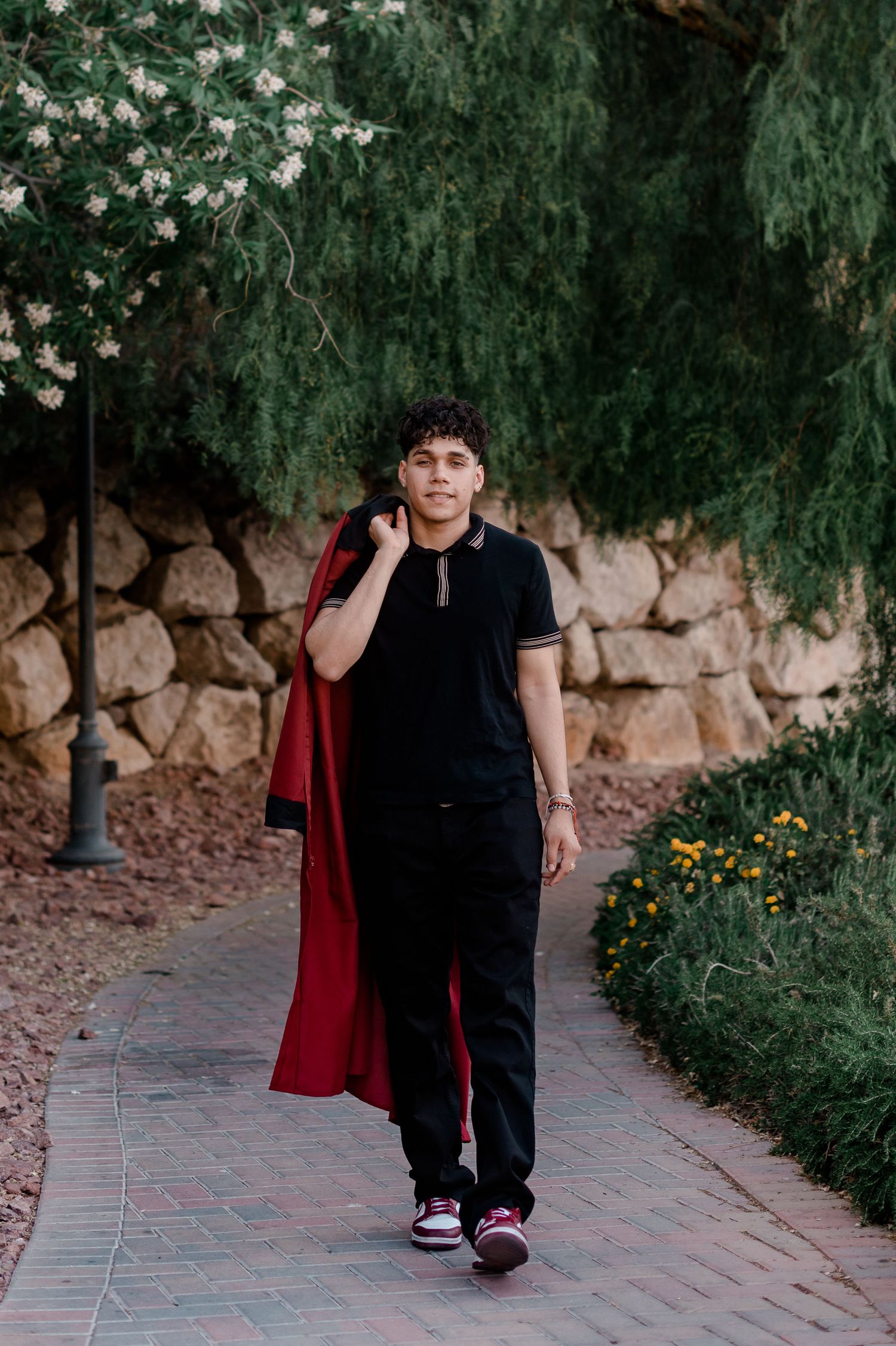 Young graduate carries red gown over shoulder walking confidently along stone-walled garden path.