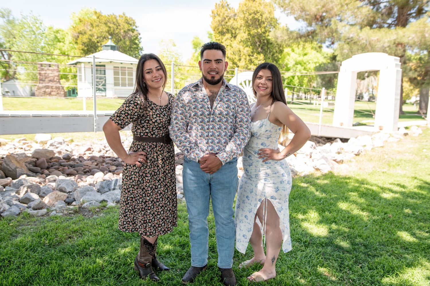 Three young adults pose together on green grass with a rocky landscape and gazebo in the background.