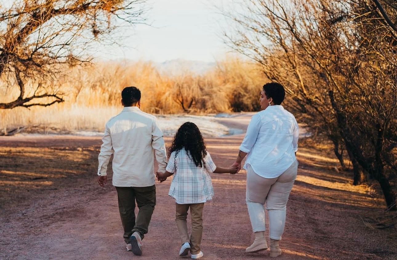 A family of three walks hand-in-hand down a dirt path surrounded by bare trees at golden hour.