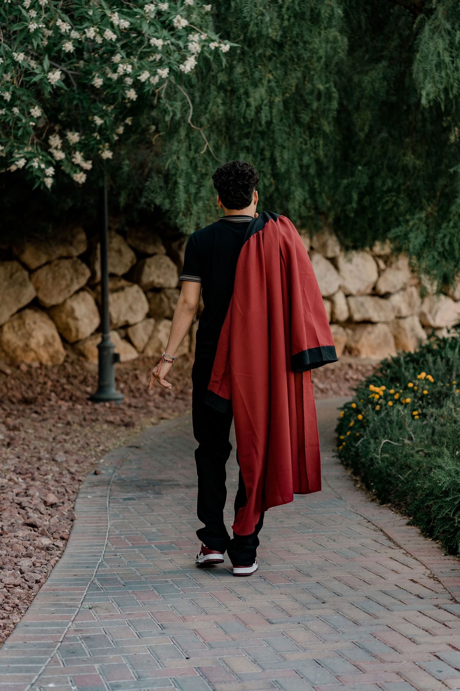 Male graduate carries red graduation gown over shoulder while walking along a scenic brick pathway.