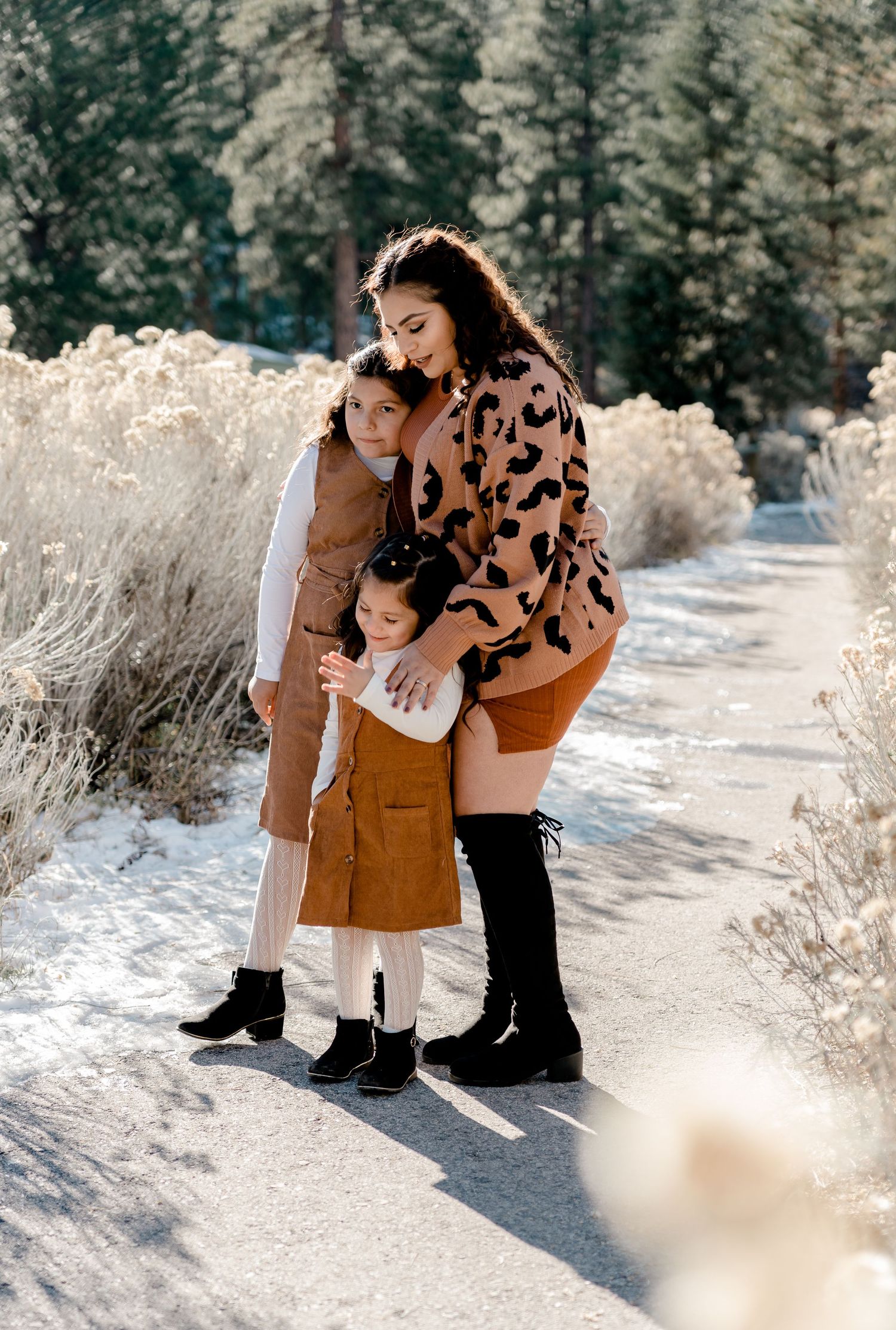 Mother hugging two young daughters on snowy winter path surrounded by frosted brush.
